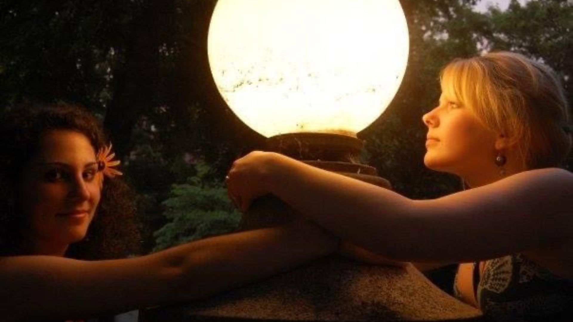 Two girls on either side of a large, round, illuminated street or house light at night with trees in background