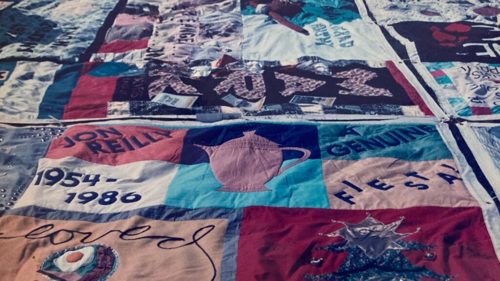 Faded quilt with titles of light brown, blue, purple, and red, with text "John Reilly" in one patch and "1954-1986" in another and "genuine" in another