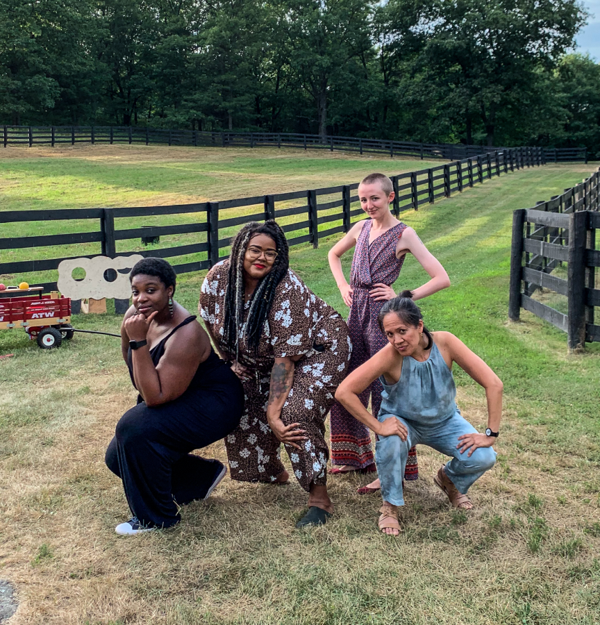 A diverse group of four writers on a grassy field pose in front of the camera. There are fences and trees in the background