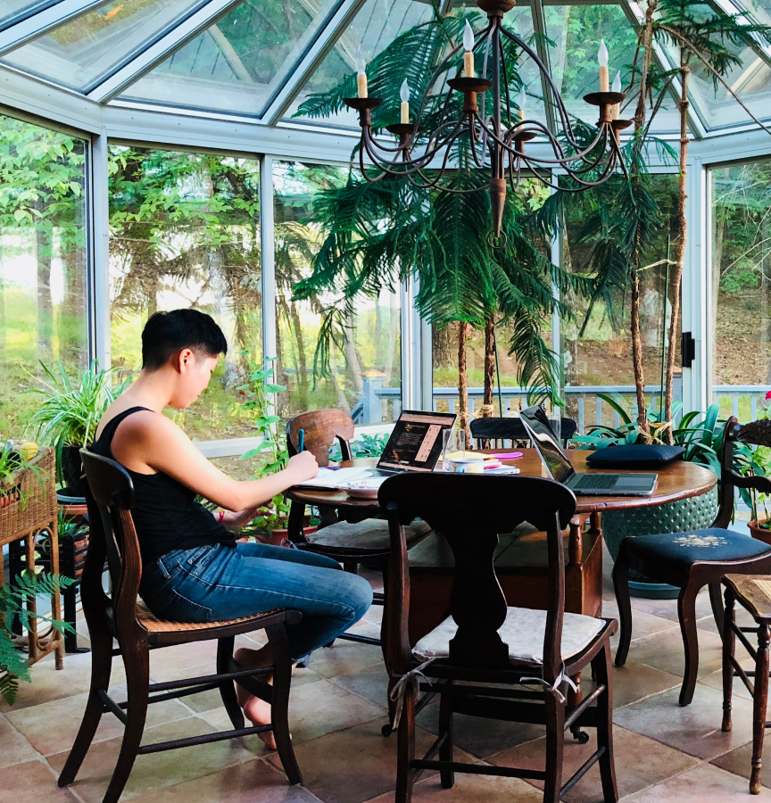 an individual writes in a sunroom on a round table surrounded by plants. There are trees outside.