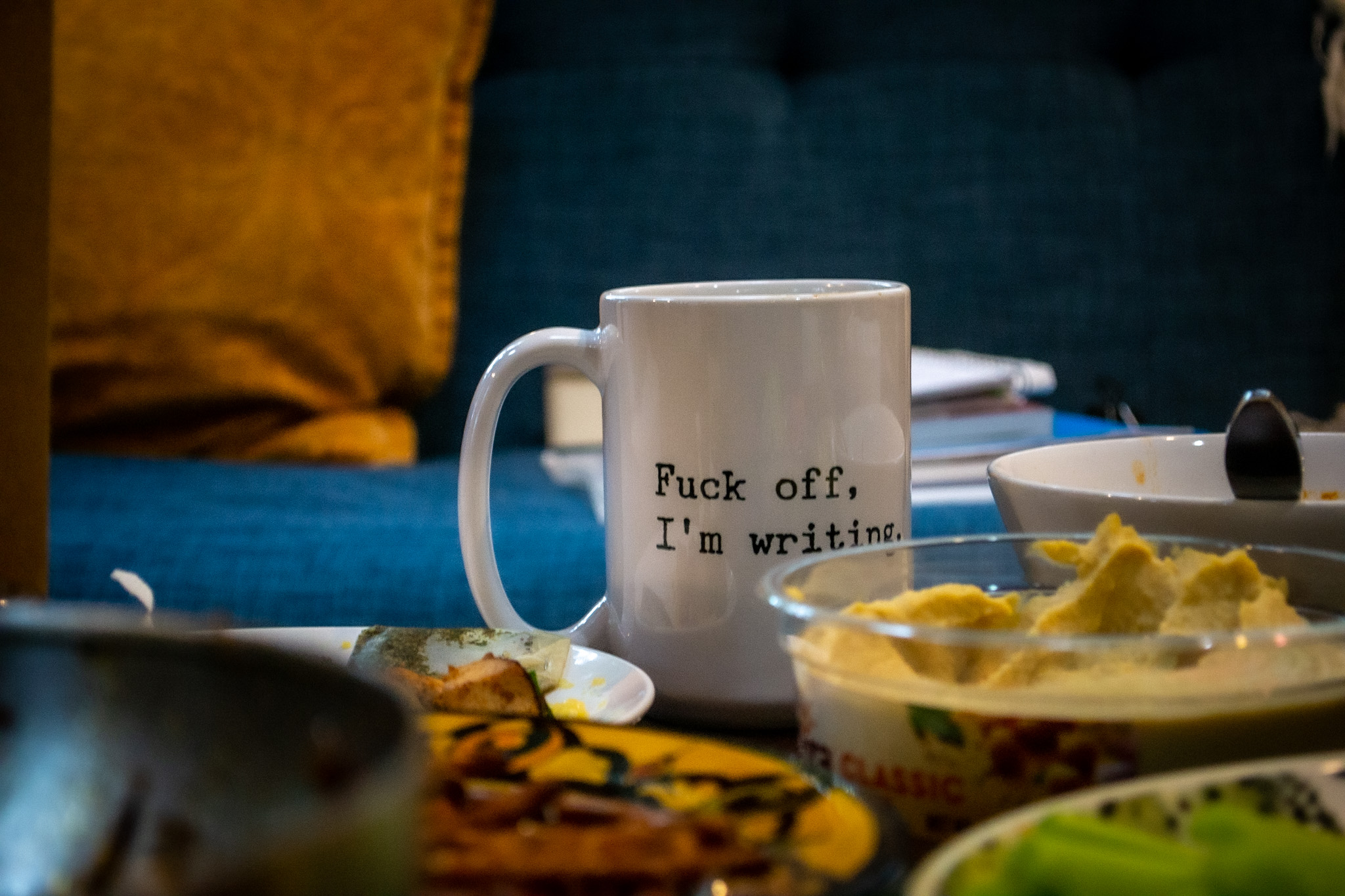 a close-up image of a mug that reads “Fuck off, I’m writing” on a table among food